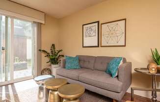 A grey couch with a blue pillow is in the middle of a room with two tables and two framed pictures on the wall.