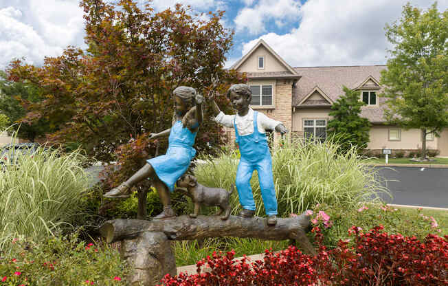 A statue of a boy and a girl sitting on a log in front of a house.