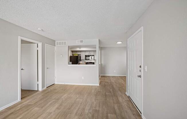 A room with white walls and wooden flooring with a kitchen visible through a doorway.