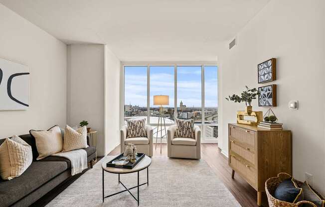 A living room with a grey couch, a coffee table, and a large window with a view of the city.
