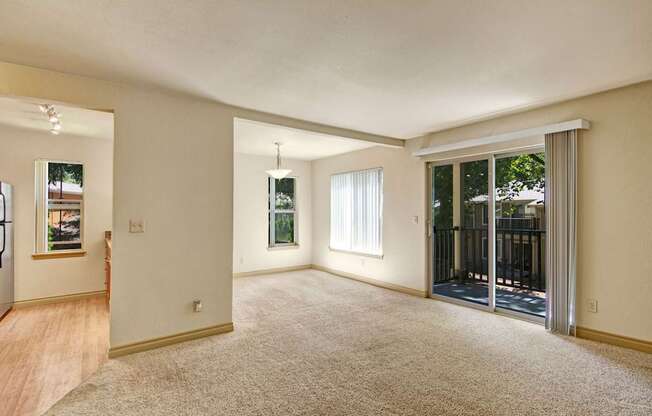A spacious open living room here at Asbury Park featuring plush carpet flooring, soft neutral walls, and a sliding glass door with vertical blinds that opens to a private balcony, complemented by an adjacent dining nook with a hanging light fixture, large windows for natural light, and a seamless flow into the kitchen area.