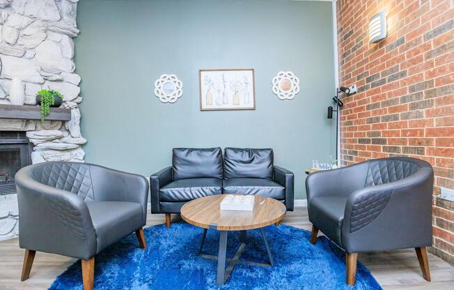 A cozy living room featuring a black leather sofa and two matching armchairs with diamond-patterned backs. In the center, there is a round wooden coffee table on a blue area rug. The walls have a mix of stone and brick accents, and framed artwork with floral motifs is displayed above the sofa.