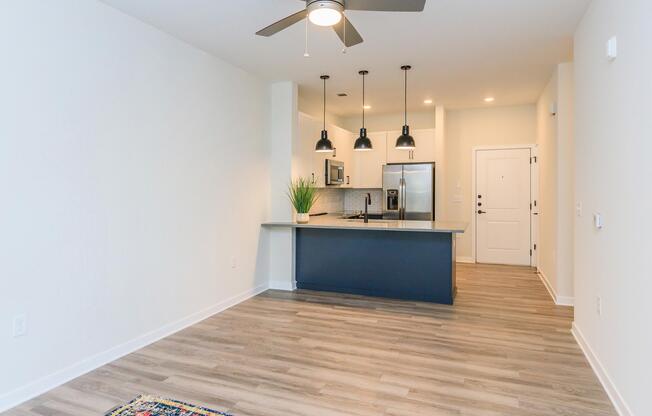 A modern kitchen and living area featuring a sleek countertop, stainless steel appliances, three pendant lights hanging above, and light wood flooring. The room has a neutral-colored wall, a door leading outside, and a decorative rug partially visible on the floor.