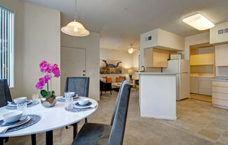 A spacious open dining and living area here at Sonoma Landing featuring a round dining table set with place settings and upholstered chairs beneath a pendant light, flowing into a cozy living room with a sofa accented by warm-toned pillows, while a breakfast bar, light cabinetry, and an in-home laundry area create a functional layout filled with natural light.