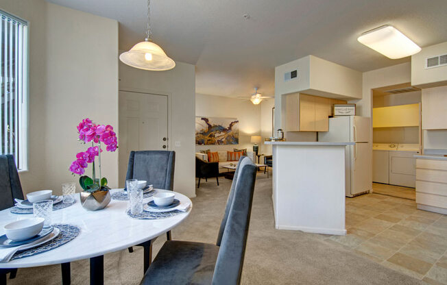 A spacious open dining and living area here at Sonoma Landing featuring a round dining table set with place settings and upholstered chairs beneath a pendant light, flowing into a cozy living room with a sofa accented by warm-toned pillows, while a breakfast bar, light cabinetry, and an in-home laundry area create a functional layout filled with natural light.