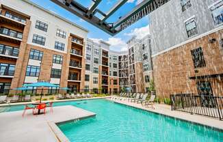 A swimming pool is surrounded by chairs and umbrellas in front of apartment buildings.