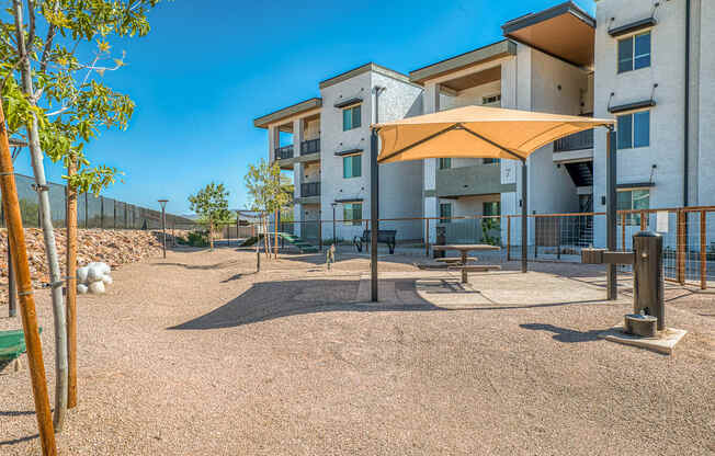 A playground area with a bench, a swing set, and a shade structure is located in front of apartment buildings.