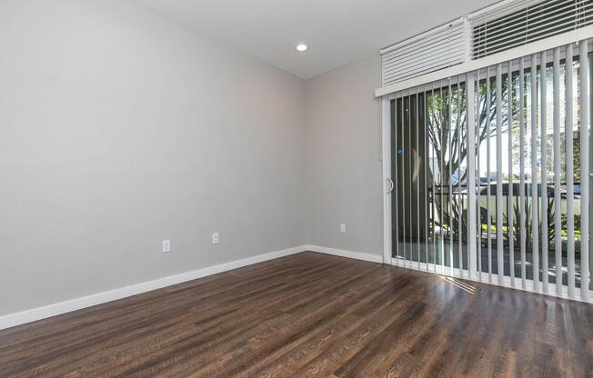 Empty room with hardwood flooring, light-colored walls, and a sliding glass door leading to an outdoor area. Natural light enters through the door, and window blinds are partially opened. The room has no furniture or decorations, creating a spacious and minimalistic feel.