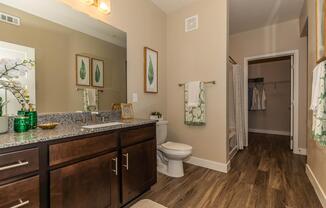 A modern bathroom featuring a double sink vanity with dark wood cabinetry, a large mirror, and decorative green plants on the counter. A white toilet is nearby, alongside towels hanging on a rack. The floor has wooden planks, and a shower area is visible in the background. Bright lighting creates a welcoming ambiance.