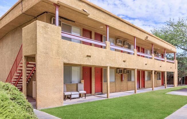 Exterior view of a two-story apartment building with a stucco facade. The building features red doors, air conditioning units, and a stairway leading to the upper level. A small seating area with two chairs is visible in front of the building, surrounded by green grass and landscaping. The sky is partly cloudy.