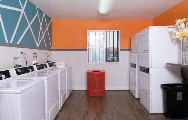 On site laundry at Fifteen 50 apartments in Las Vegas with four washers and dryers, red trash can, and modern orange, blue, and white walls.