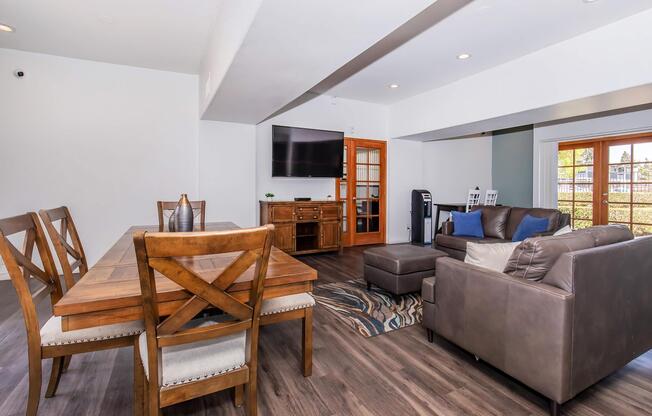 An inviting modern living space featuring a wooden dining table with chairs, a comfortable brown sofa, an ottoman, and a TV mounted on the wall. Large windows allow natural light, and there are decorative elements like a rug and plants, creating a warm and cozy atmosphere.