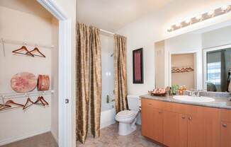 A bathroom here at Landing at Mansfield with a wood-tone vanity, gray countertop, large mirror, bright vanity lights, tan patterned shower curtain, tub-shower combo, and an open closet with hanging racks.