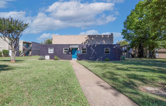 A laundry center with a blue door is surrounded by a grassy area and trees at The Drake in Bossier City, LA