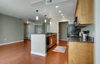 A bright open kitchen and living area here at Bridges with warm wood flooring, light wood cabinets, dark countertops, black appliances, a center island with pendant lights, and an open layout leading to nearby rooms.