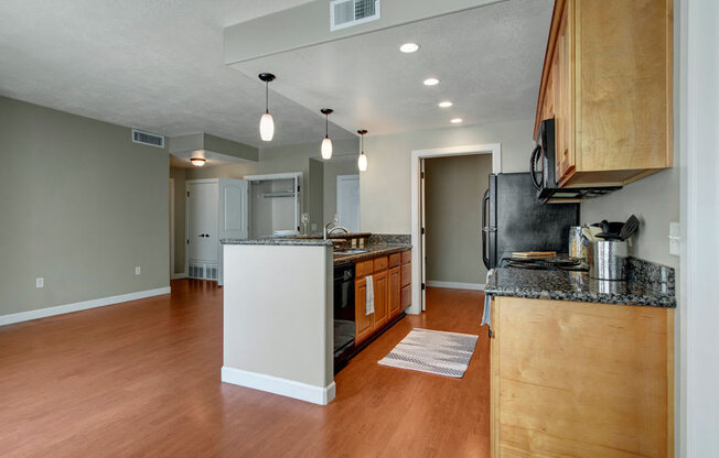 A bright open kitchen and living area here at Bridges with warm wood flooring, light wood cabinets, dark countertops, black appliances, a center island with pendant lights, and an open layout leading to nearby rooms.