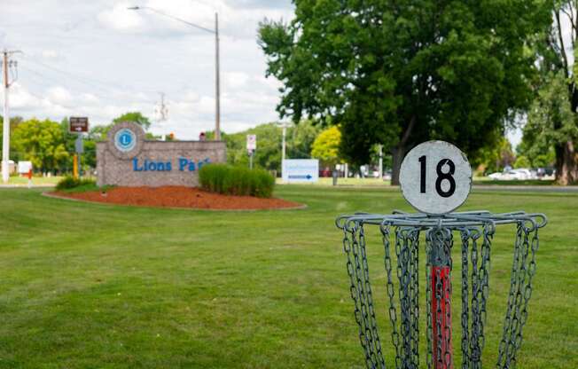 A disc golf basket with the number 18 on it is in the foreground of a grassy area with a sign that says Lions Park in the background.