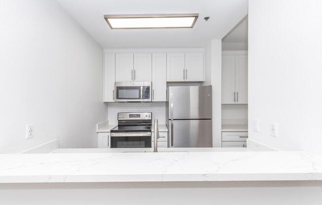A modern kitchen featuring white cabinetry, a stainless steel refrigerator, and an oven with a microwave above it. The countertops are light-colored with a marble-like design. The kitchen is well-lit, with a bright atmosphere and an open design that connects to another room.