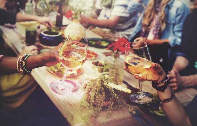 Stock photograph of people sitting around a table and eating. Two wine glasses are being clinked in the foreground.