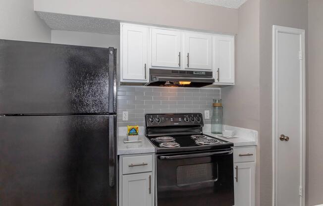 A modern kitchen featuring black appliances, including a refrigerator and an electric stove. The white cabinetry contrasts with the dark stove and fridge, while a gray tile backsplash adds a sleek touch. Countertops are light-colored, and a door on the right leads to another space.