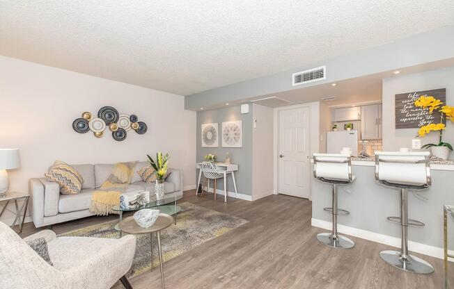 A modern living room featuring a light gray sofa with yellow and gray accents, a glass coffee table, and art on the walls. Adjacent is a small dining area with white chairs and a kitchen in the background, showcasing sleek cabinets and decor highlighted by a yellow flower arrangement.