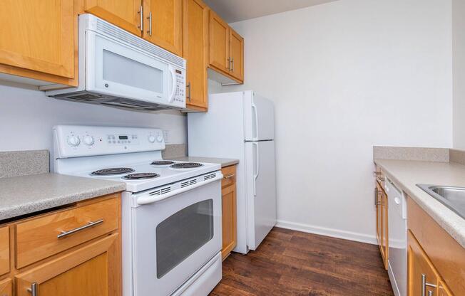 A modern kitchen featuring light wood cabinets, a white stove with an oven, a microwave above, and a refrigerator. The countertops are a light beige, and there's a stainless steel sink. The flooring is a dark wood laminate, creating a bright and clean look.