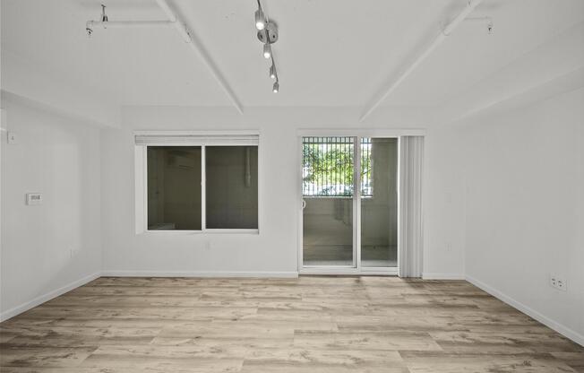 Empty modern living room with light-colored walls and wooden flooring. A large window on the left side offers natural light, while a sliding glass door leads to a balcony or patio. There are exposed beams on the ceiling and track lighting overhead, creating a bright and spacious atmosphere.
