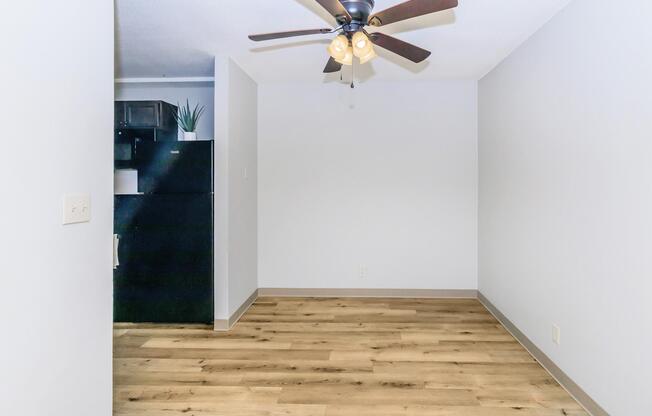 Empty room with light-colored walls and wooden flooring. A ceiling fan with light fixtures is visible, and at the back, there is a black refrigerator. The space is well-lit, creating a clean and open atmosphere.