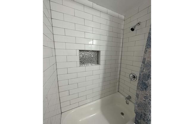 A modern bathroom shower area featuring white tiled walls, a built-in shelf with mosaic tiles, and a bathtub. The showerhead is mounted on the wall, and a decorative shower curtain partially covers the bathtub. The space is well-lit and designed for functionality.