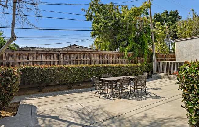 A patio with a table and chairs is surrounded by a hedge and a wooden fence.