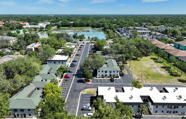 Scenic aerial view of a charming apartment complex surrounded by lush greenery and serene water views, showcasing spacious parking areas and well-maintained buildings.