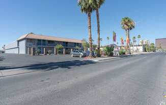 A street view with an apartment building, palm trees, and a car.