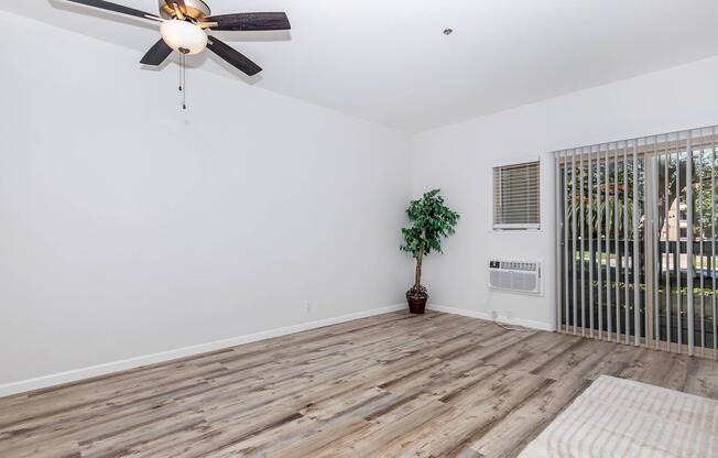 A spacious, empty interior room featuring light wood floors, a ceiling fan, a large window with vertical blinds, and an air conditioning unit. A small indoor plant is placed in a pot, adding a touch of greenery to the otherwise minimalistic space. The walls are painted white.