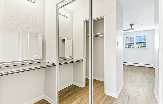 Elegant apartment interior featuring a spacious walk-in closet, stylish mirrored doors, and sleek wooden flooring leading to a bright, airy room with large windows and modern ceiling fan.