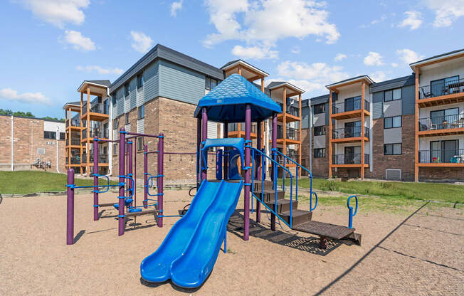A playground with a blue slide and a play structure in front of apartment buildings.