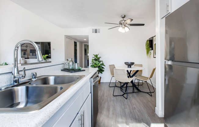 Modern kitchen featuring a stainless steel sink, stylish faucet, and gray cabinetry. A dining area with a round table and four chairs is visible, along with plants for decor. The space has a bright, airy feel, illuminated by a ceiling fan and natural light.