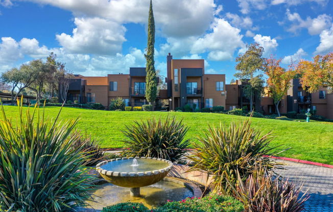 a fountain in the middle of a grassy area with apartment buildings in the background