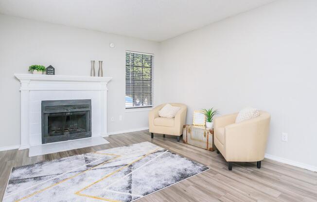 A cozy living room featuring a white fireplace, two beige armchairs, and a decorative area rug with geometric patterns. A small table and a plant add a touch of greenery. Large window with blinds allows natural light to brighten the space. Walls are painted in a light color, creating an inviting atmosphere.