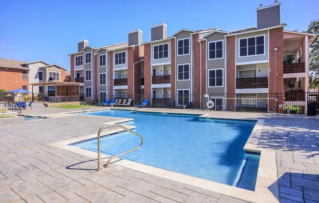 A well-maintained outdoor swimming pool surrounded by a concrete patio, with lounge chairs nearby. In the background, there are modern multi-story apartment buildings featuring balconies and large windows, set against a clear blue sky.
