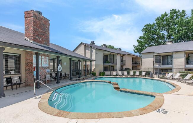 Swimming pool with a heart-shaped design, surrounded by lounge chairs. There are brick and stucco apartment buildings in the background, along with trees and a clear blue sky overhead.