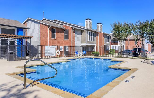 A clean swimming pool surrounded by a paved area, located in front of a two-story brick apartment building. The pool features a metal ladder and is enclosed by a black fence. Visible amenities include lounge chairs and a safety buoy. The sky is clear and blue, indicating a sunny day.