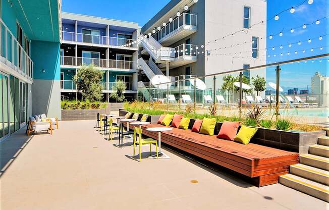 A patio with a long bench and chairs is surrounded by buildings.