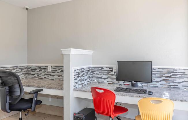 A modern workspace featuring a grey wall, a granite countertop, and a desktop computer with a keyboard and monitor. Two office chairs are positioned in front, one black and one yellow, alongside a red chair. There is a decorative backsplash of small grey tiles, adding a stylish touch to the area.