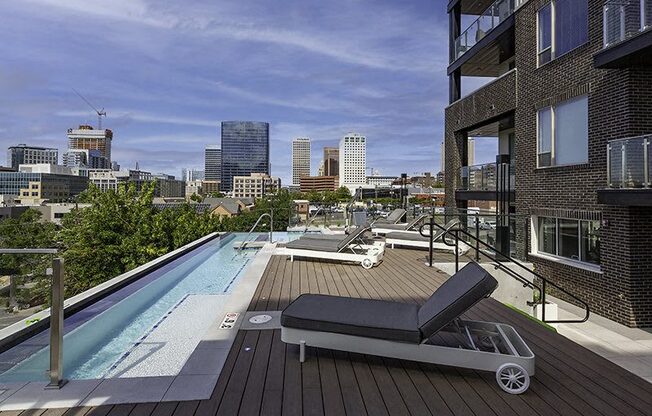 A rooftop pool with a lounge chair and a hot tub with a city skyline in the background.