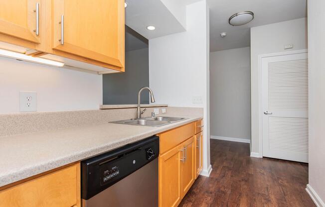 A modern kitchen featuring wooden cabinets, a stainless steel sink, and a black dishwasher. The countertop is light-colored, and there is a well-lit space with a view toward a hallway that has a closed white door at the end. The flooring is dark wood, enhancing the contemporary feel of the space.