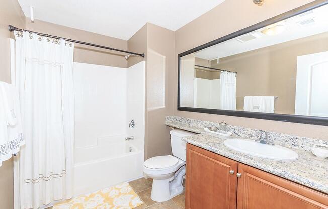 A well-lit bathroom featuring a bathtub with a shower, beige walls, and a large mirror above a granite countertop. The countertop has a sink and toiletries. There's a towel rack with white towels, and a decorative yellow mat on the floor adds a pop of color.