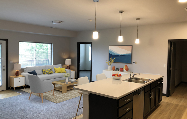A modern, bright open-concept living space featuring a kitchen island with a sink, pendant lighting, and fruit. The living area has a gray couch with colorful throw pillows, a light rug, and a small coffee table. A painting hangs on the wall, and large windows provide natural light.