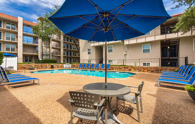 A pool area with lounge chairs, patio tables and chairs with blue umbrellas and stainless steel BBQ in front of apartment buildings at Princeton Court Apartments in the Vickery Midtown neighborhood of Dallas, TX.