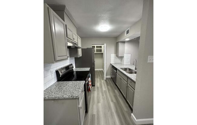 An interior view of a modern kitchen featuring gray cabinets, a granite countertop, stainless steel appliances, and a tile backsplash. The kitchen includes a stove, refrigerator, and sink, with ample natural light. The flooring is light wood laminate, creating a clean and contemporary look.