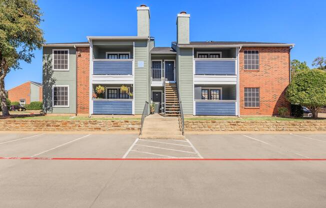 A two-story apartment building with a mix of brick and blue siding. The front porch has stairs leading up to the second floor, with planters on either side. There are several parking spots in front, and the sky is clear and blue, indicating a sunny day.